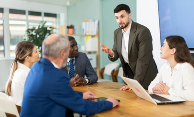 Obraz premium Man stands in front of business colleagues, tells report, proves vision of situation, defends way to solve problems. People are sitting at table, and participant is standing up and took floor. .
