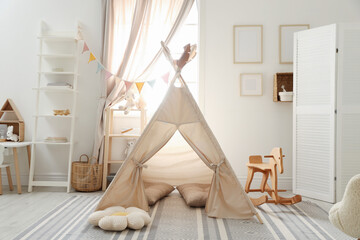 Stylish toy wigwam, furniture, pillows and decor near window in playroom. Interior design © New Africa