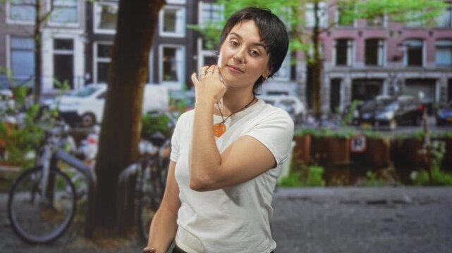 Woman touching chin while wearing orange pendant necklace on a street by a canal with bicycles and parked cars, casual white tee; serene confidence.