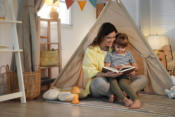 Mother and her daughter reading book near toy wigwam at home © New Africa