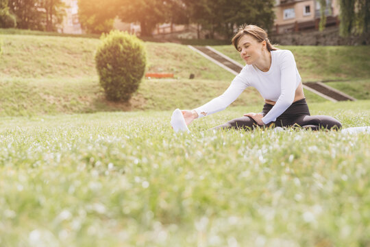Woman practicing yoga outdoors, stretching leg and touching toes on a yoga mat in green park grass