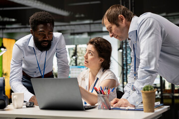 Obraz premium Diverse corporate employees in office workspace review financial data and investment reports on laptops. Professional woman conducts project briefing with male colleagues, analyzing company statistics
