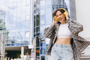 Fototapeta premium Young woman enjoying music with yellow headphones, smiling happily in city street, urban lifestyle