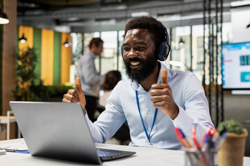Obraz premium Black man sits at desk, using laptop for video conference with business associates and giving thumbs up for successful presentation. Smiling employee showing positive gesture to team in virtual