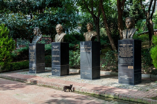 A stray cat strolls past bronze busts of world historical figures in a leafy Istanbul park garden, Kadikoy