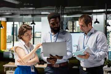 Fototapeta premium Black man and two white colleagues working together on financial reports and business investment strategies. Multiethnic office team standing and reviewing company performance on laptop.