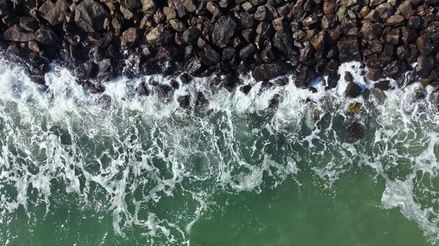 Waves roll forward and hit the rocky shoreline. Water splashes against stones as foam builds up. The scene shows the meeting of sea and land during daylight hours.
