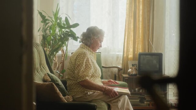 Side view of senior woman with grey hair looking through photo album reminiscing bittersweet memories of youth then leaning back on couch in retro living room