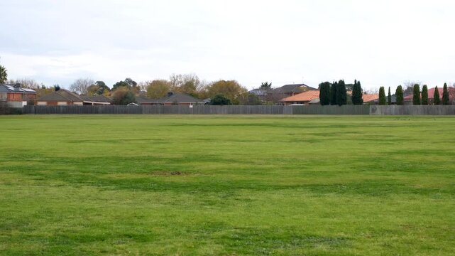 Pan shot of an open grassy field in a suburban neighborhood in Hoppers Crossing, Melbourne, Australia, with houses in the background under an overcast sky.  Wide spacious green space.