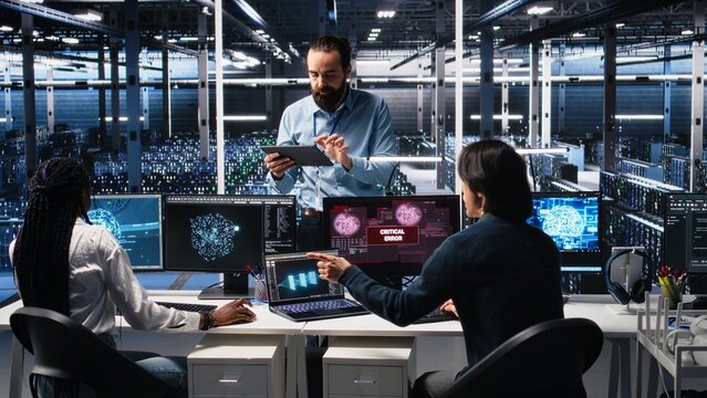 System administrators in data center struggle with computer bugs while using AI. Frustrated server farm coworkers doing brainstorming to fix system errors while using artificial intelligence, camera A