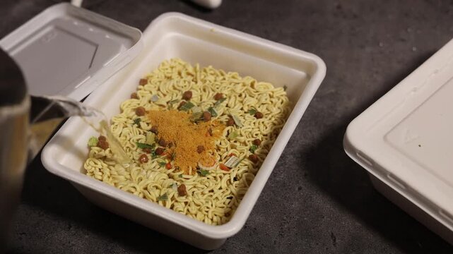 Cook pours hot boiling water from electric kettle, over quick cooking instant egg curly noodles, ramen with spices, herbs in plastic packaging for soup and Asian plates. Concept of healthy fast food