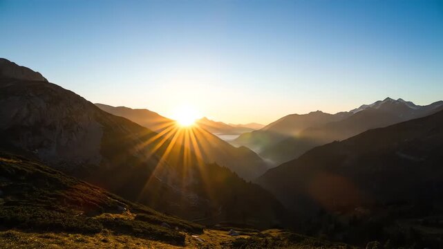 A scenic vista shows the sun setting over mountain peaks. Valley below fills with clouds, as sky shifts color, from blue to yellow