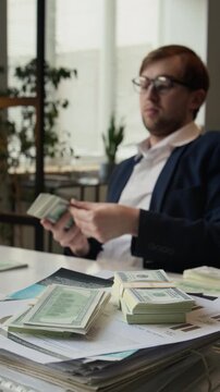 Vertical selective focus view of smug gen Z entrepreneur in formalwear counting hundred-dollar bills at workplace