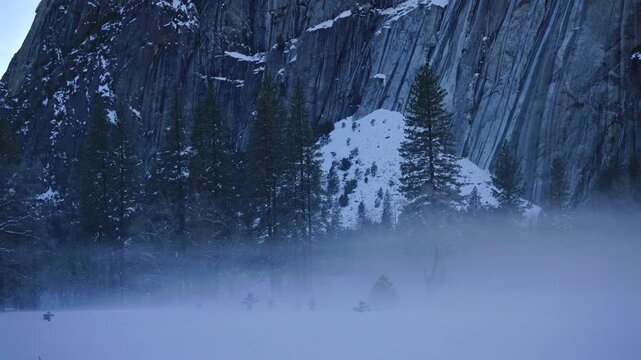 Snowy Yosemite National Park valley at dusk, mist rising across meadow, towering granite cliff face dusted with snow, solitary pine silhouette, icy blue light, hushed atmosphere evoking solitude