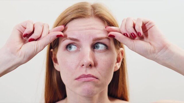 Red-haired Woman examines her forehead lines while pulling skin between her fingers in a well-lit indoor space at home