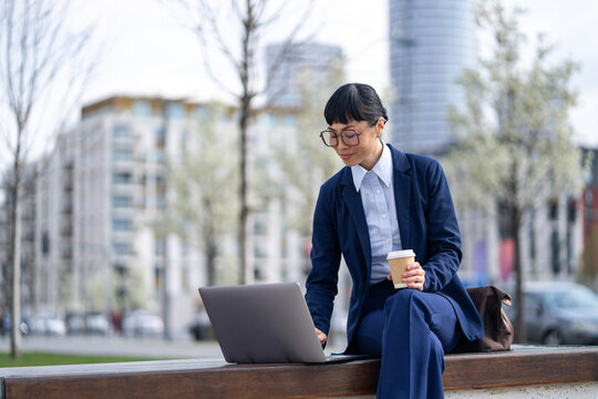 Businesswoman working on laptop outdoors with coffee, urban background
