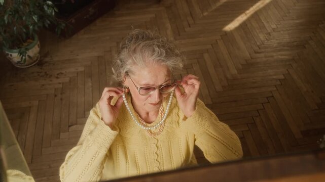 High angle shot of senior woman with white hair putting on vintage pear necklace while sitting at vanity table in bedroom, slow motion