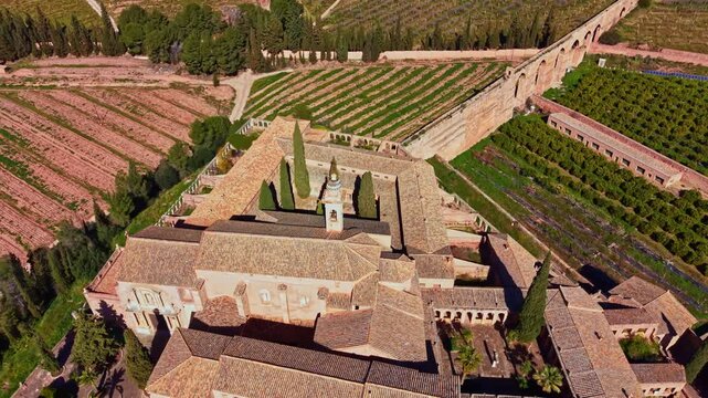 A large building stands in the center with multiple sections. Surrounding it are green fields, vineyards, and trees. The scene shows a bright sky overhead.