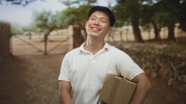 Man holding cardboard box smiling and laughing on a dirt street by a gate under trees, gesturing with free hand; joy hope.