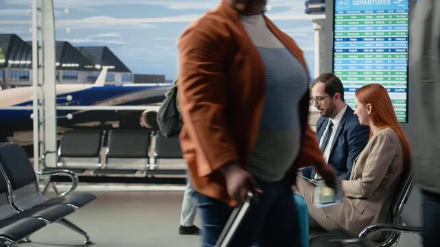 Business colleagues on corporate travel delegation in airport waiting area, discussing and waiting for boarding plane near the gate. Passengers checking flight schedule with carry-on suitcases.