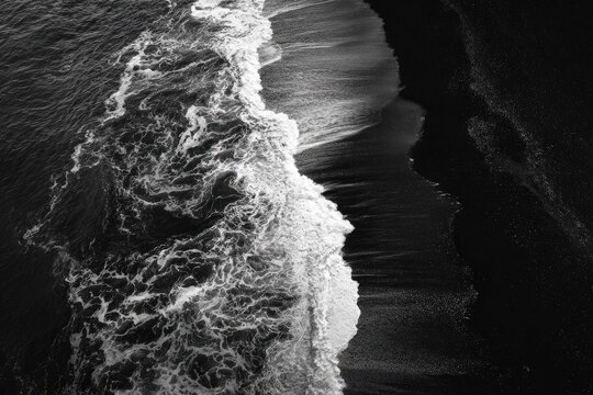 Aerial, monochrome view of ocean waves crashing onto dark sand. Contrast between foam and shore