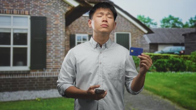 Man holding creditcard and smartphone, eyes closed and head tilted up while pointing card outward in front of building; digital transaction uncertainty.