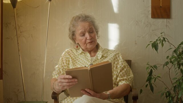 Medium shot of impressed elderly woman with grey hair thinking plot over reading novel in hardcover book while sitting in armchair in retro apartment