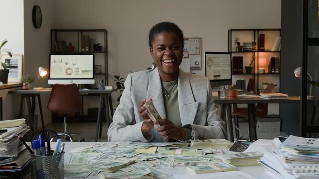 Zoom out portrait of happy Black young woman wearing grey jacket looking at camera holding bundle of dollar bills and smelling them while sitting at white table cluttered with money