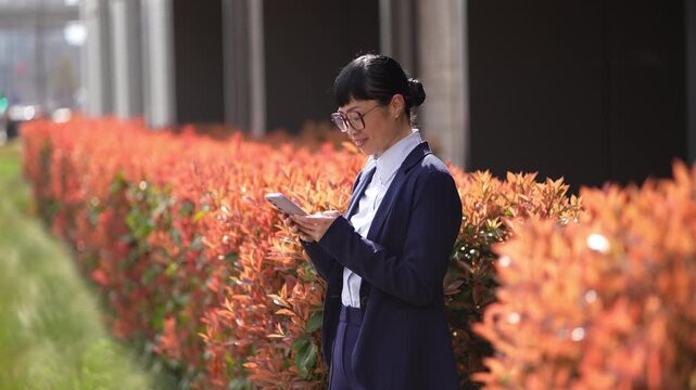 Businesswoman in glasses checks her phone while walking past vibrant orange bushes