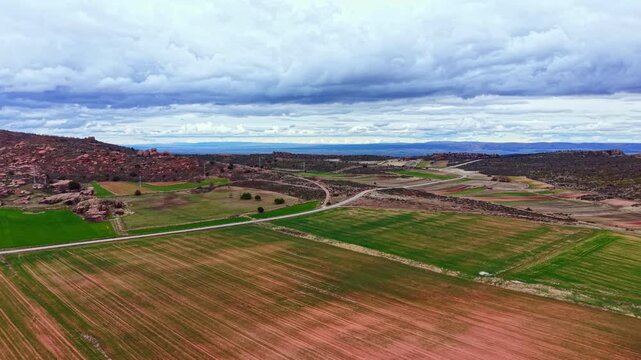 Green fields stretch across the land with dirt roads visible. Clouds cover the sky, creating a dramatic look. The rural area shows signs of agricultural use with varying soil colors.