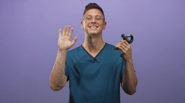 Physiotherapist man in teal scrubs waves left hand holding handgrip exerciser in studio; friendly rehabilitation support.