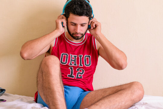 Man listening to music sitting on the bed, holding headset, relaxing, holding it. Handsome bearded man with a fit body, red team shirt and blue underwear. Legs and arms visible.