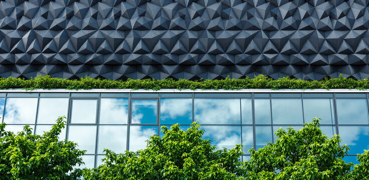 Contemporary architecture featuring dark triangular panels, glass windows reflecting sky, lush greenery on roof and foreground, urban design, eco friendly structure.