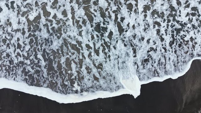 Waves roll onto a dark sandy beach under a bright sky. The ocean water interacts with the land, creating white foam at the edges. This scene shows the constant movement of the sea.