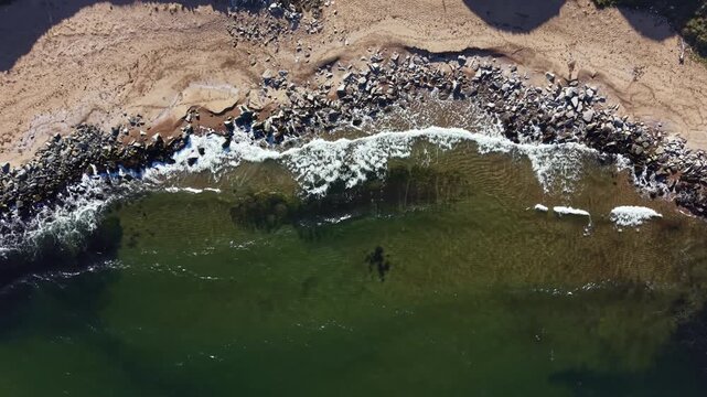 A person is swimming near a beach while waves crash on the shore. Rocks outline the water's edge. This scene captures a sunny midday with clear water.