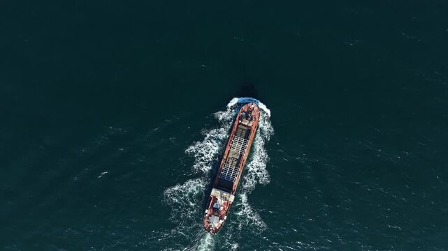 A cargo ship moves through the open water under bright sunlight. Waves surround the vessel as it travels, creating a visible path on the water's surface.