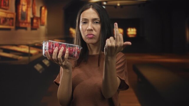 Hispanic woman holds clear container of strawberries and shows middle finger in art gallery; defiance attitude rebellion.