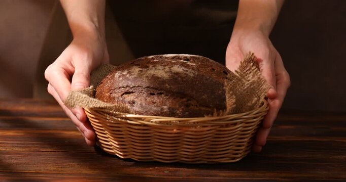 Woman with loaf of fresh rye bread in wicker basket at wooden table, closeup