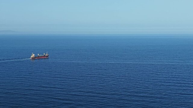 A cargo ship travels through deep blue ocean waters. The sky is clear with no visible clouds. The coastline can be seen far away. The water surface appears smooth and still.