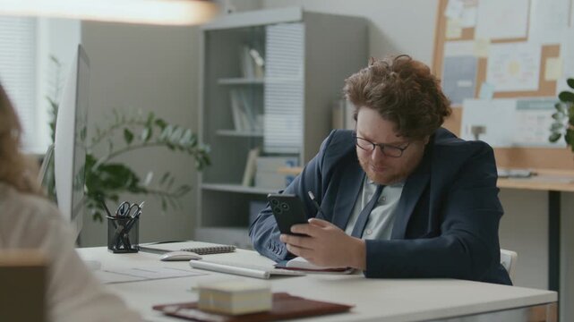 Young businessman sitting at desk in open office, using smartphone and taking notes, then typing on computer during workday