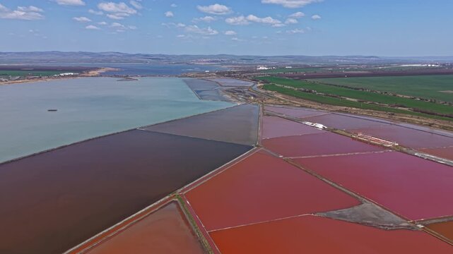 Groups of salt ponds create a patchwork of colors under a clear sky. Fields and a water body are visible in the background. This scene shows nature and human activity.