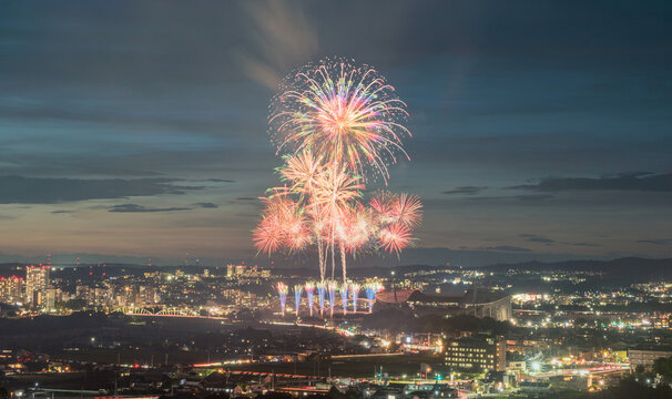 Brilliant multicolored fireworks burst into the twilight sky over a lively cityscape in Aichi, Japan. The summer festival atmosphere is enhanced by glowing lights, and a scenic urban backdrop.