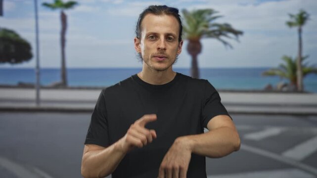 Young man with long hair in black t shirt pointing finger to wrist on street by the ocean and palm trees; time checking impatience.