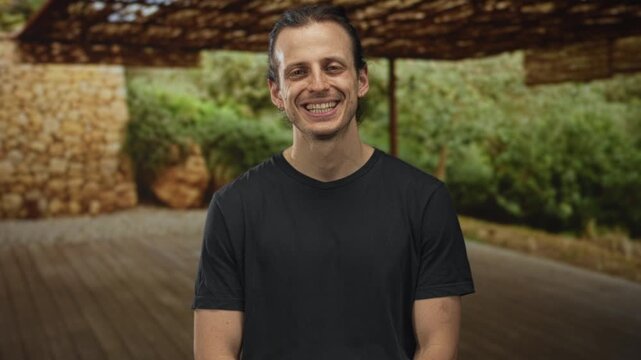 Man smiling and seated on a wooden deck in a forest pavilion under a woven canopy; serenity ease openness.