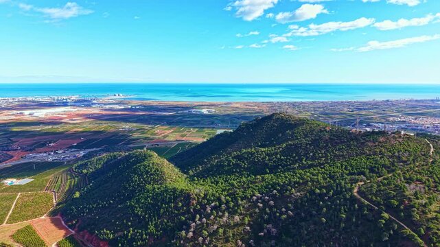 Aerial view shows green hills beside flat land and fields reaching towards the coast. The sea is visible in the background under a clear blue sky.