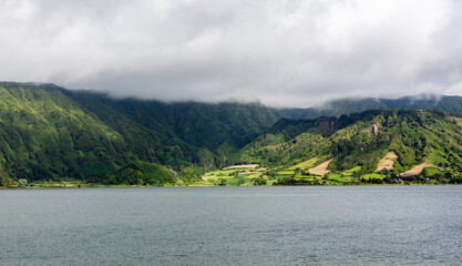 Fototapeta premium Crater lake at Sete Cidades, San Miguel, Azores. Volcanic caldera lake surrounded by lush green hills. Cloudy sky over serene water landscape. Located in Portugal.