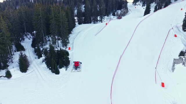 A snow groomer works on a ski slope in a mountain area. This activity takes place in winter with clear weather. Trees surround the ski area, creating a scenic view.