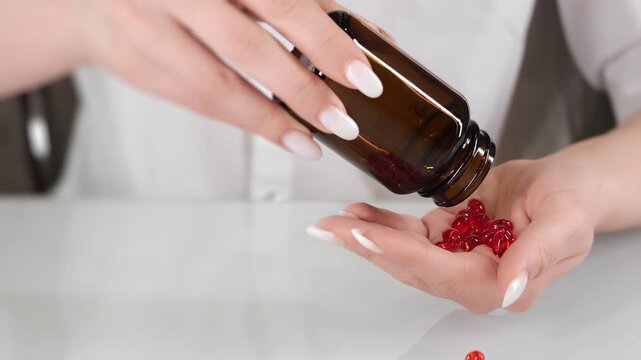 Close up of female hands pouring red capsules from glass bottle into palm on clean background representing supplement intake healthcare and wellness routine.