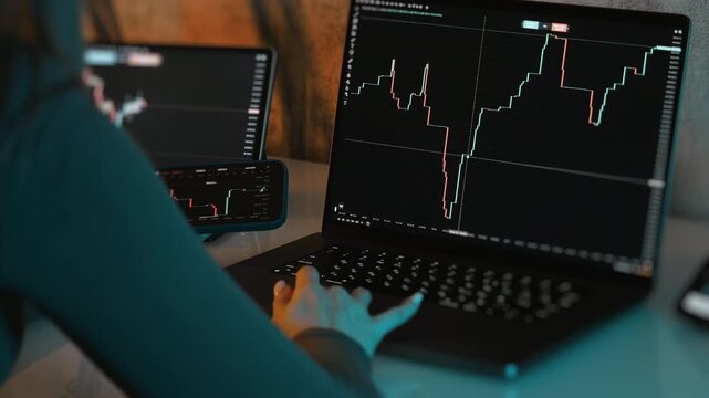 Over the shoulder view of trader using laptop and smartphone with stock market charts in modern workspace representing financial investment and online trading concept.