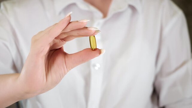 Close up of female hand holding single omega capsule between fingers on clean background representing vitamin intake healthcare and wellness concept.
